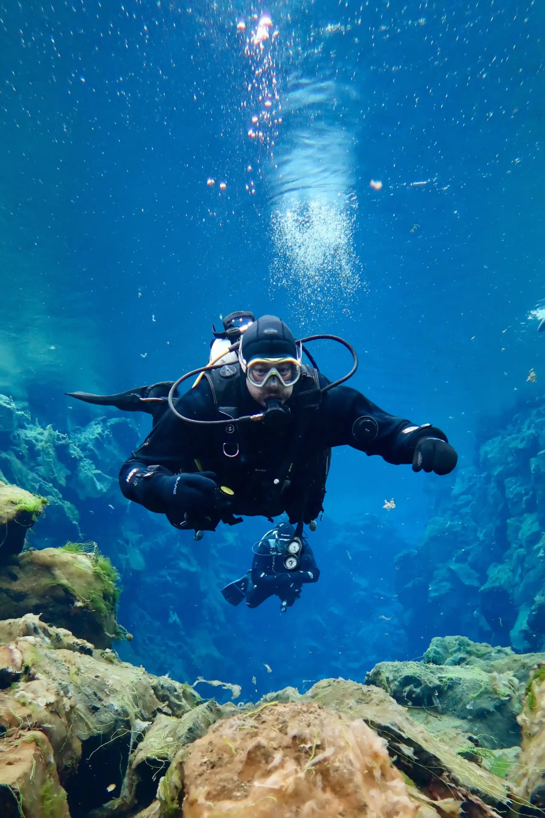 A scuba diver in a drysuit swims toward the camera between rocks in clear blue water