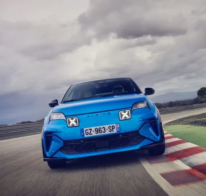 A blue Alpine A290 drives toward the camera on a race track, past red and white curbs